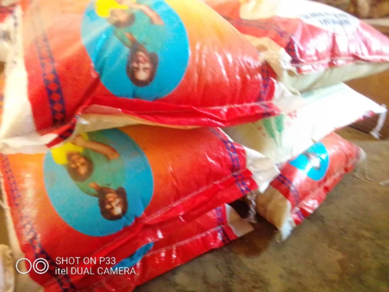 Bags of rice stacked on a table, featuring red and blue packaging with a circular logo. The scene conveys a sense of supply and abundance.