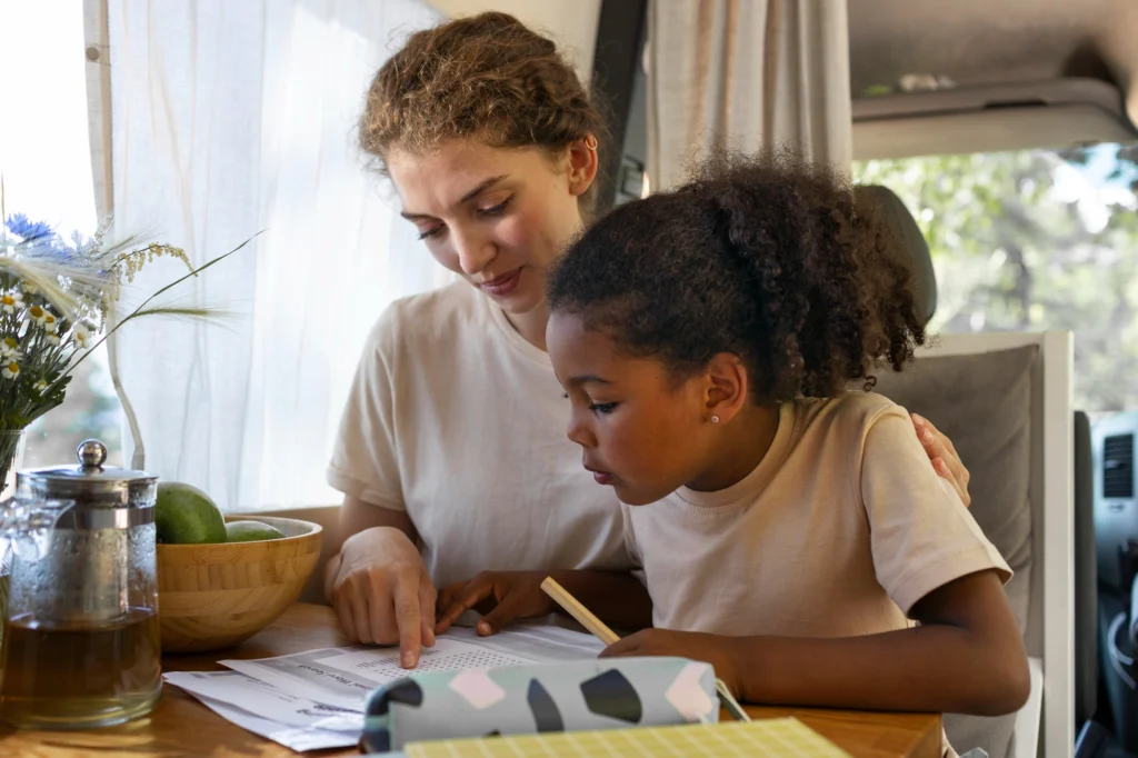 A mentor helps an orphaned or vulnerable child in Cameroon with schoolwork. The child has an arm around the mentor, showcasing a warm, safe environment. A wooden pencil case and paperwork are visible.