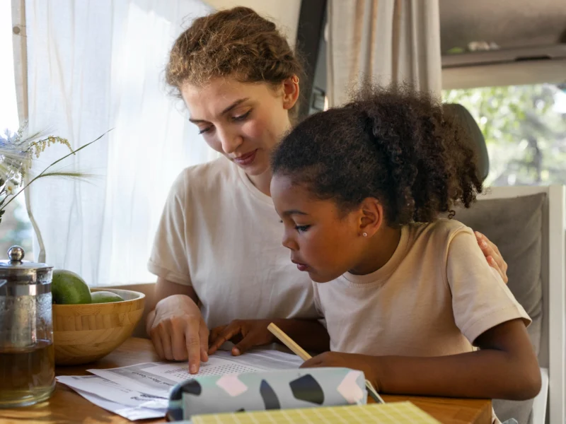 A mentor helps an orphaned or vulnerable child in Cameroon with schoolwork. The child has an arm around the mentor, showcasing a warm, safe environment. A wooden pencil case and paperwork are visible.