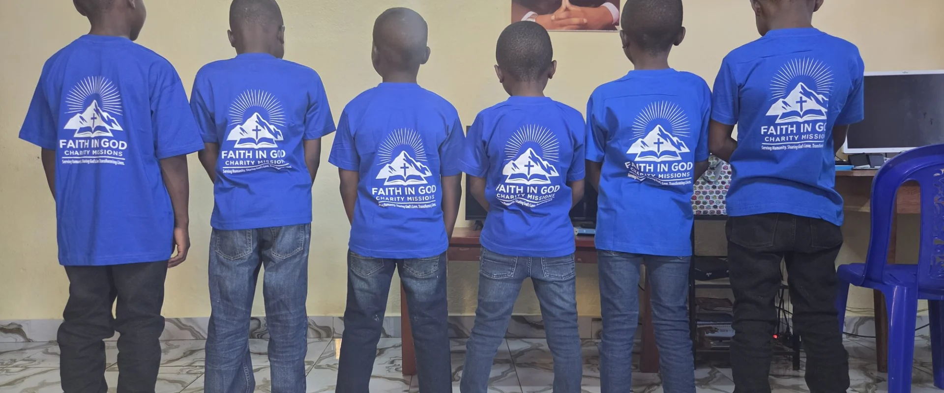 Children in blue Faith in God Charity Missions shirts standing together indoors.