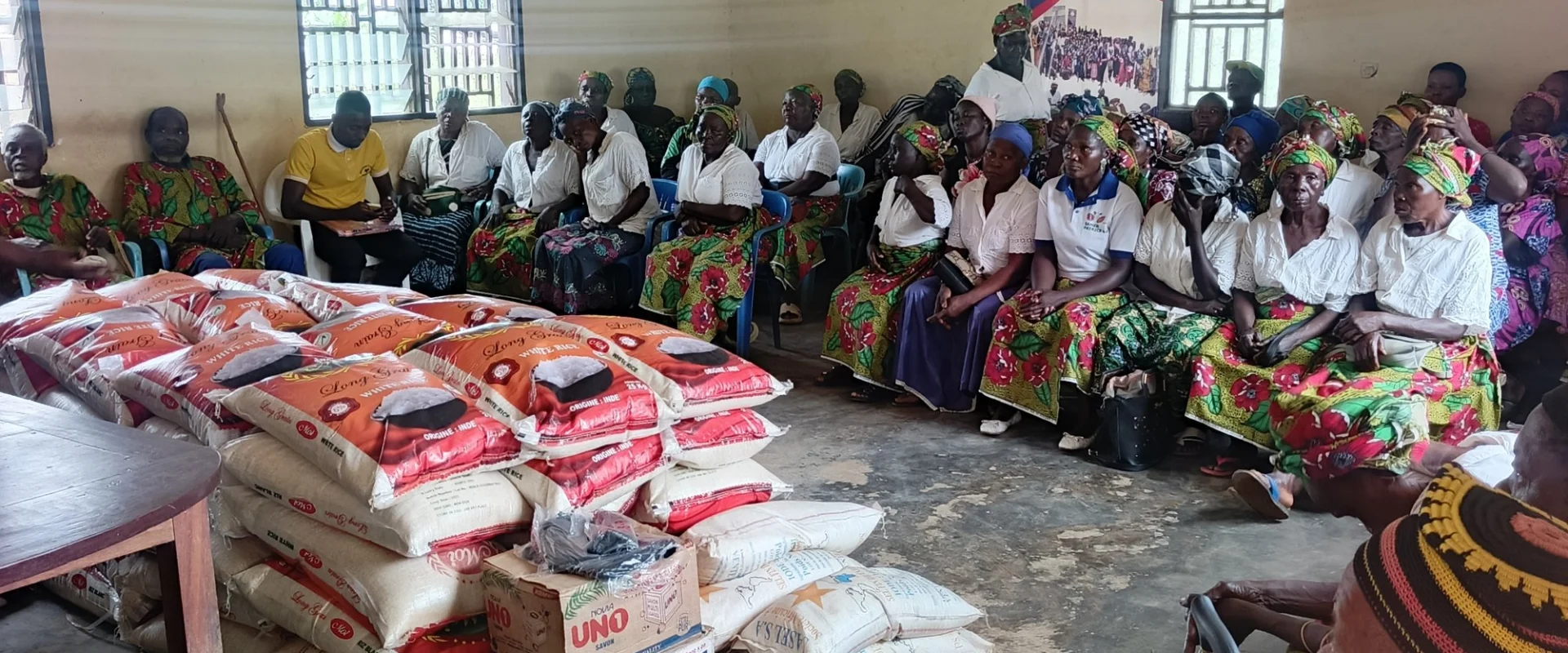 Sacks of rice and other supplies stacked ready for distribution, with recipients seated behind them.