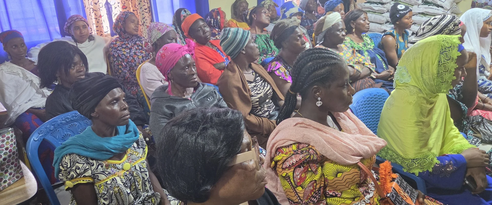 Women seated together at a community distribution, with supplies stacked at the back of the room.