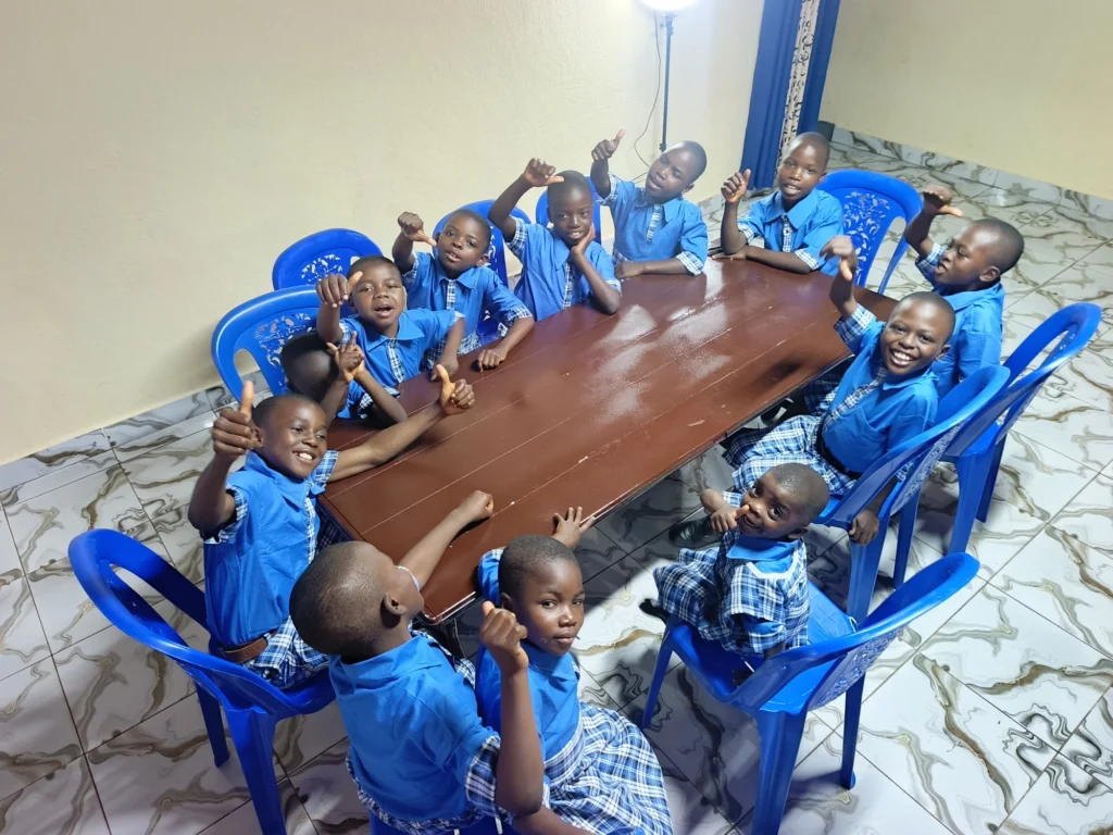 Children in blue uniforms around a table, raising their hands and giving a thumbs up.