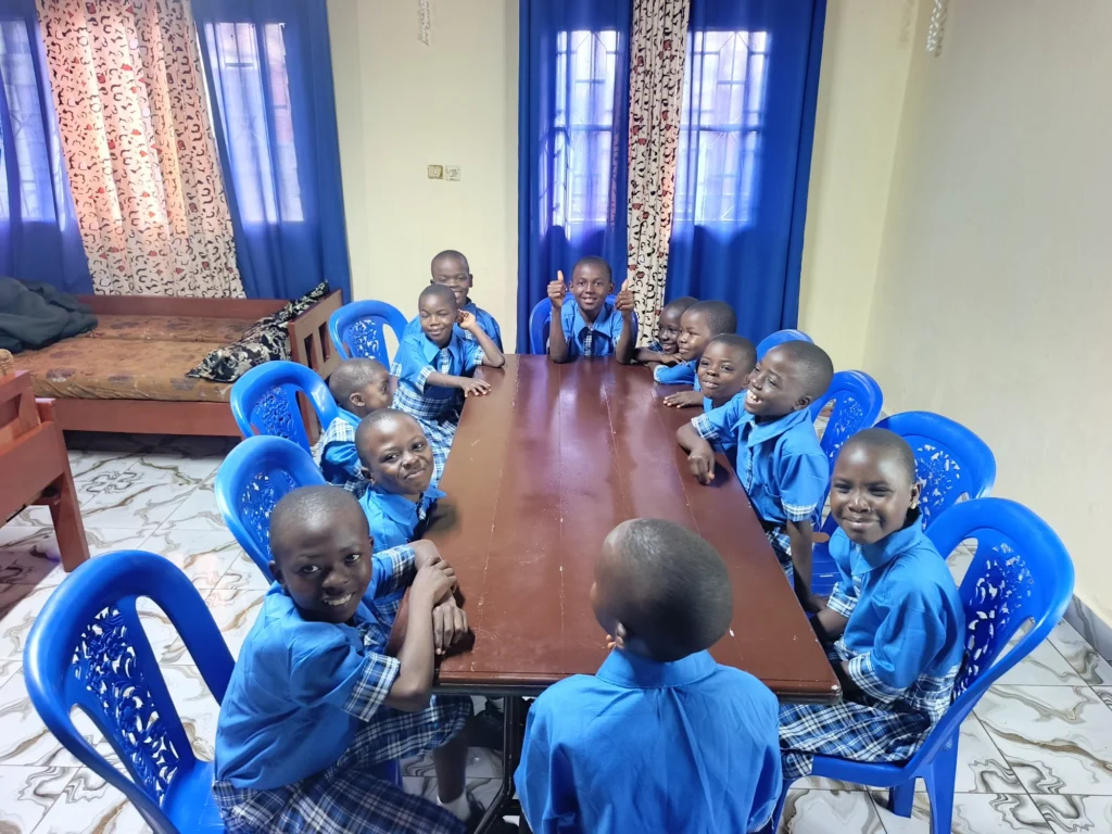 Children in blue school uniforms seated around a table at the FIGCM orphanage, smiling and giving a thumbs up.