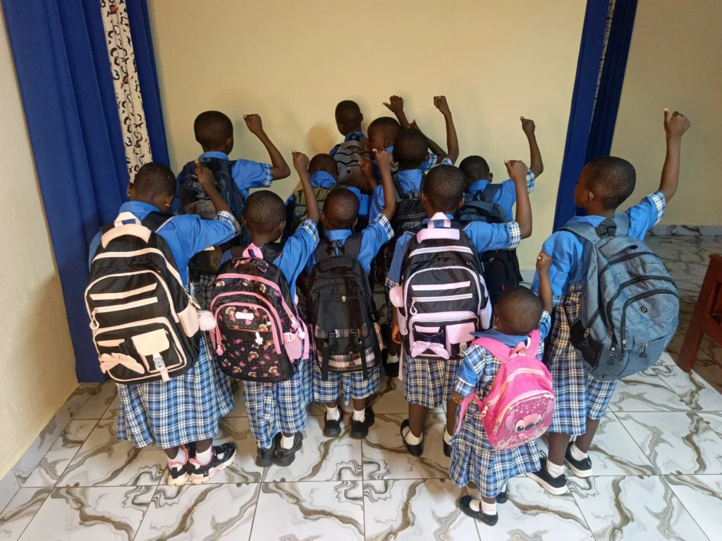 Children in blue uniforms with backpacks raising their arms as they head out for school.